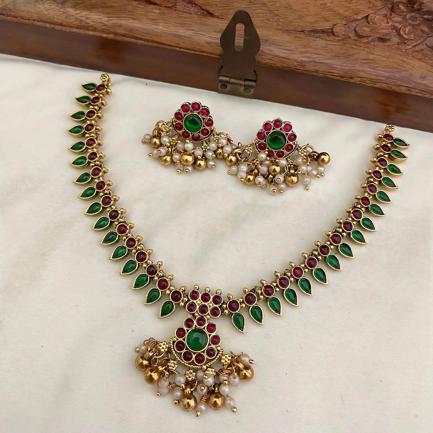 Gold necklace with green and red stones on a white cushion with a wooden box in the background