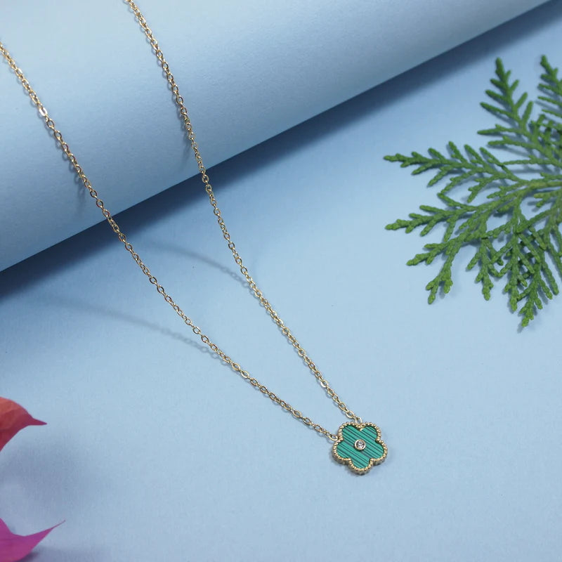 Gold necklace with a green pendant on a light blue background with green leaves.