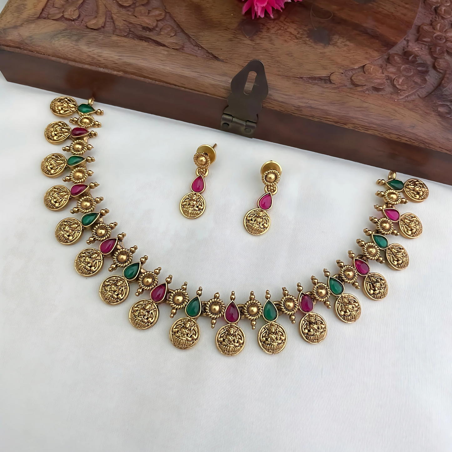 Gold necklace with green and red stones and matching earrings on a white surface with a wooden box in the background.