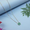 Gold necklace with a green pendant on a light blue background with green leaves.