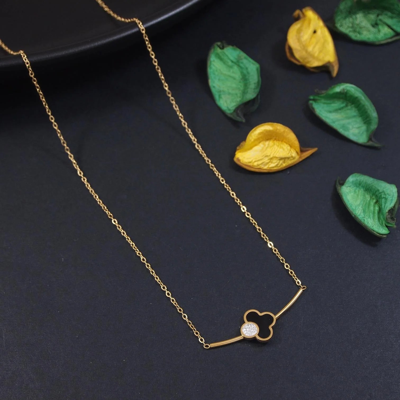 Gold necklace with a clover-shaped pendant on a dark surface with leaves.